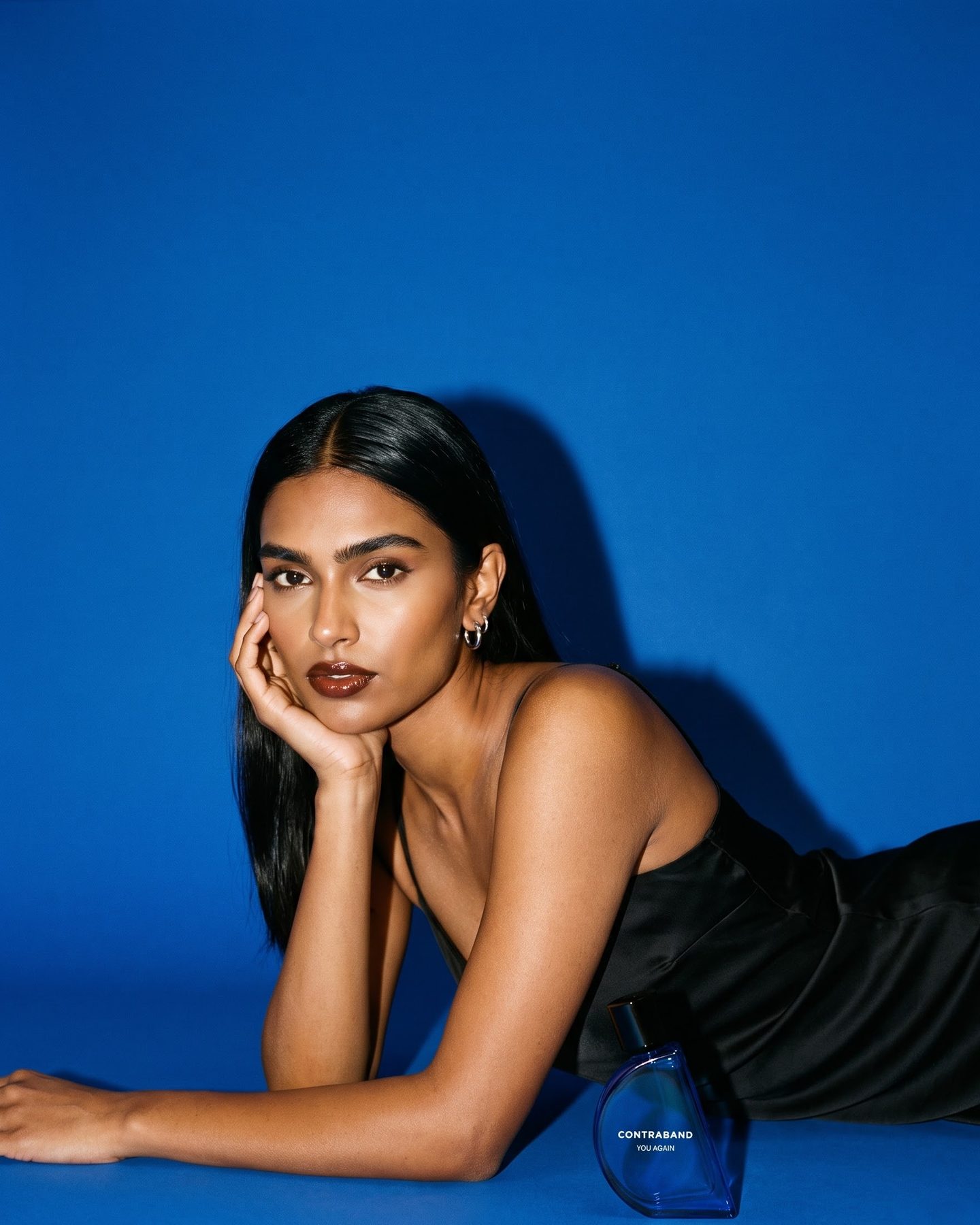 On a cobalt-blue studio set, a woman in a black satin slip lies propped on her forearms, chin in hand, a cobalt Contraband bottle beside her elbow.