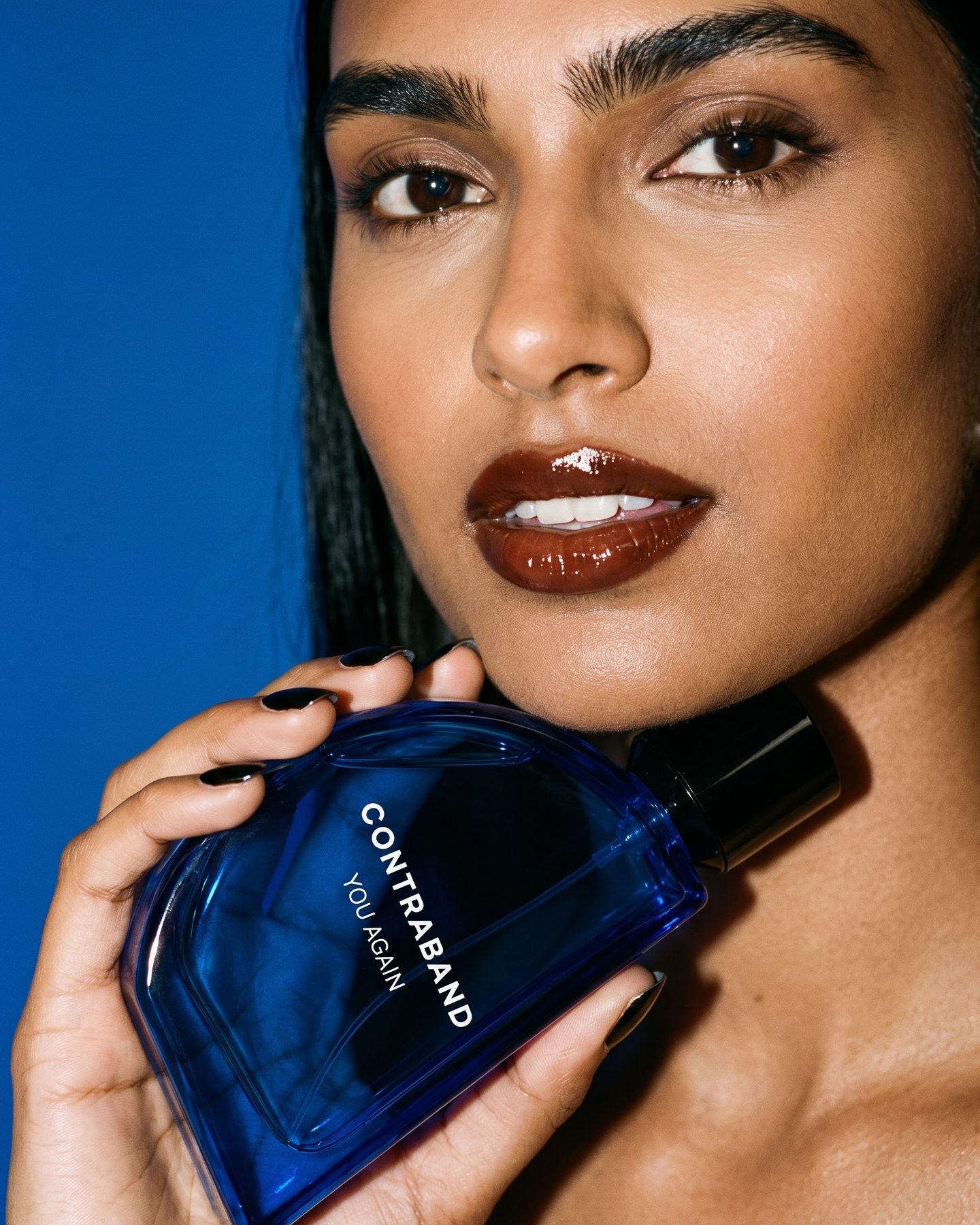 Beauty close-up of the woman with glossy cherry-brown lips, holding the Contraband You Again bottle against her jaw, black nails.