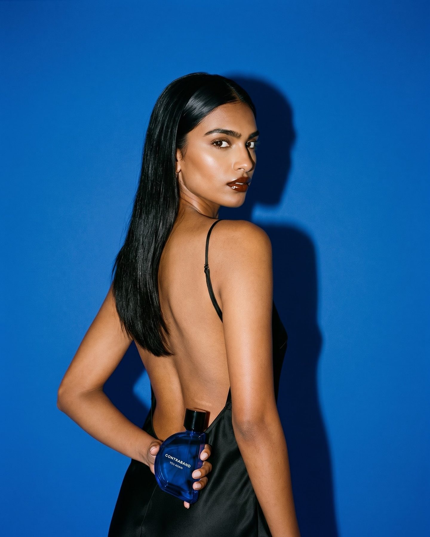 The woman stands with her back to the camera in an open-back black slip dress, looking over her shoulder, the bottle held behind her back.