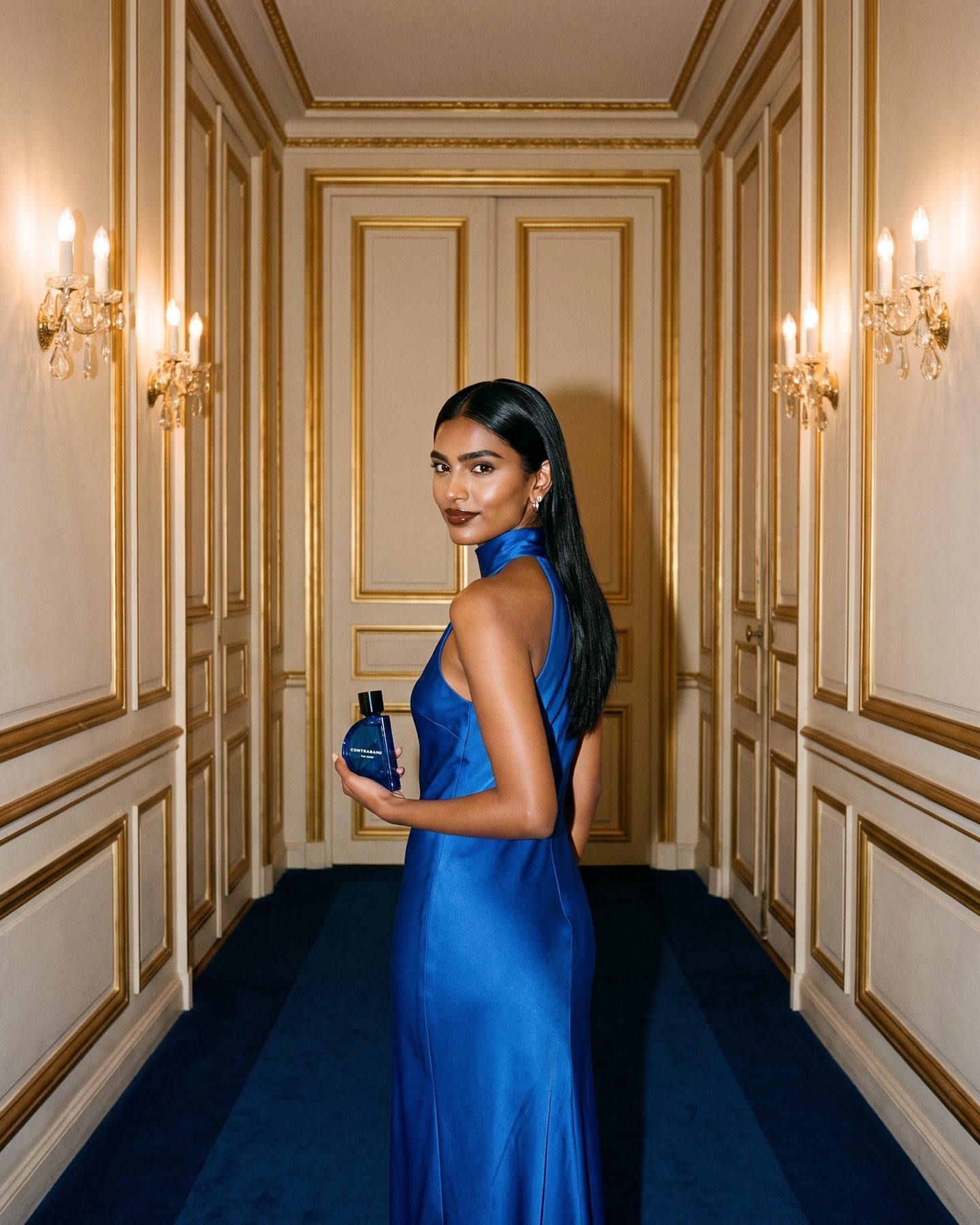 In a gilded Parisian hotel corridor with a cobalt carpet, the woman in a cobalt silk gown turns back toward the camera, holding the bottle.