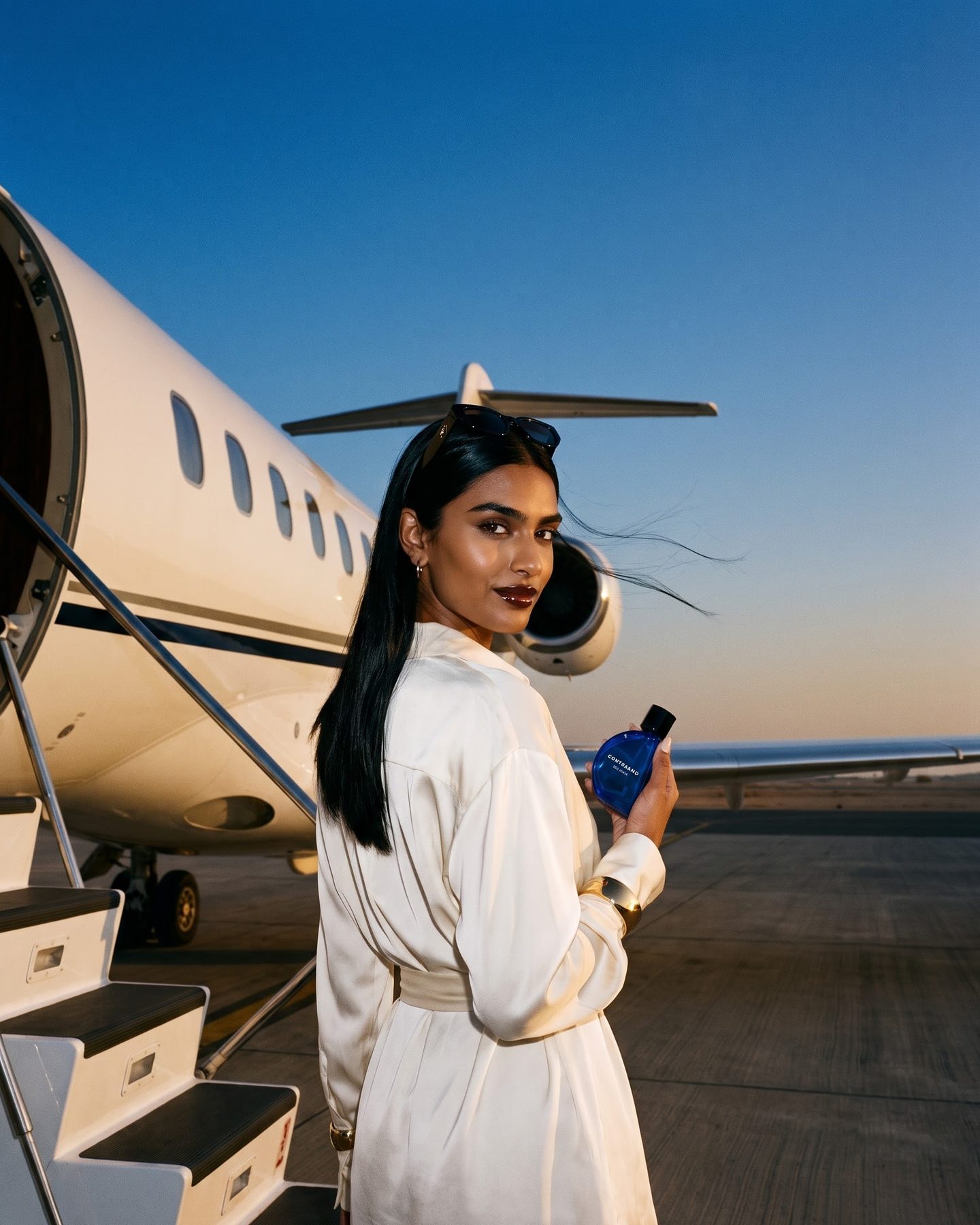At blue hour on a desert airfield, the woman in an ivory silk shirt dress stands at the foot of a private jet's airstairs, looking back, bottle in hand.
