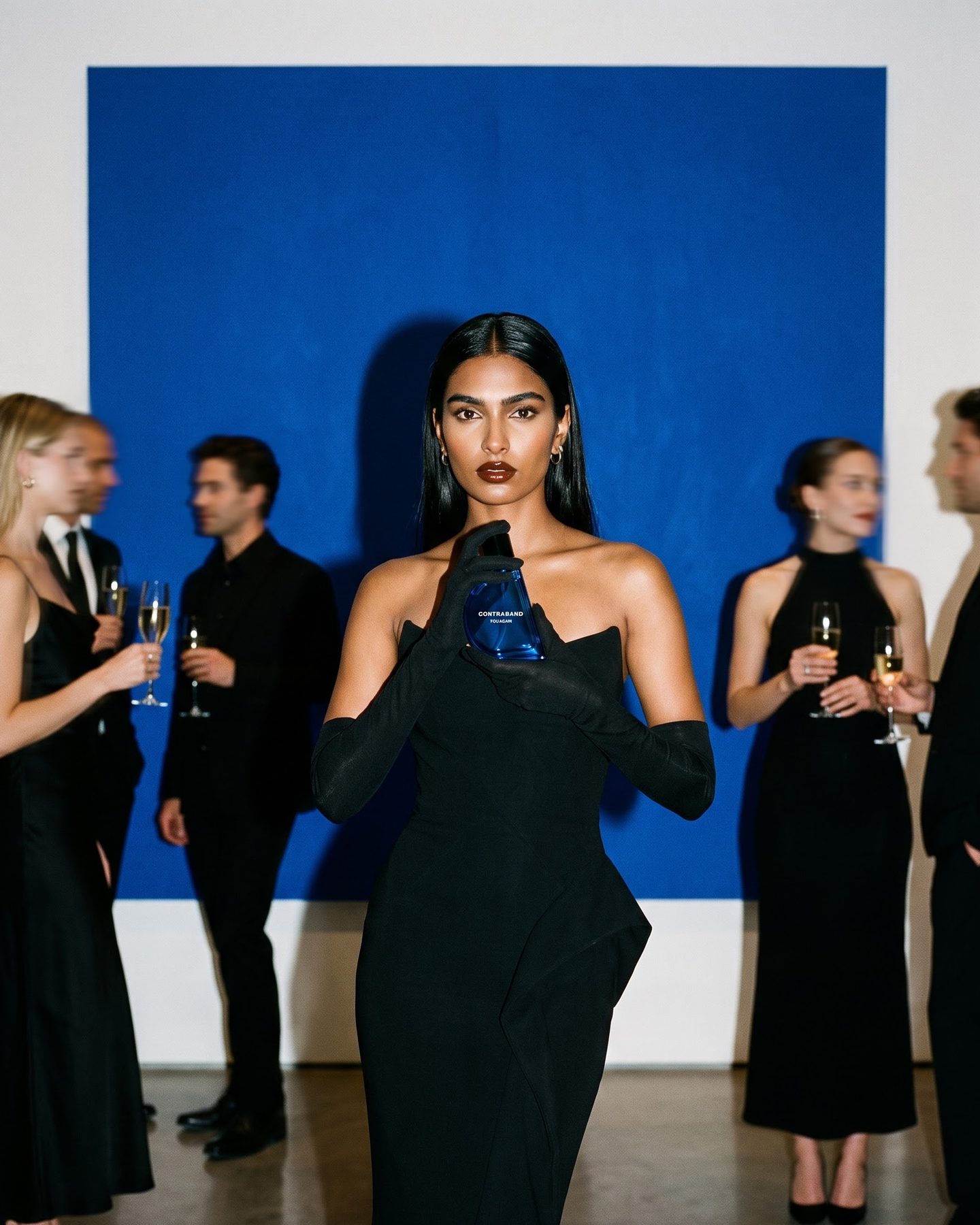 At a gallery opening in front of a huge cobalt canvas, the woman in a black strapless gown and opera gloves holds the bottle while guests blur around her.
