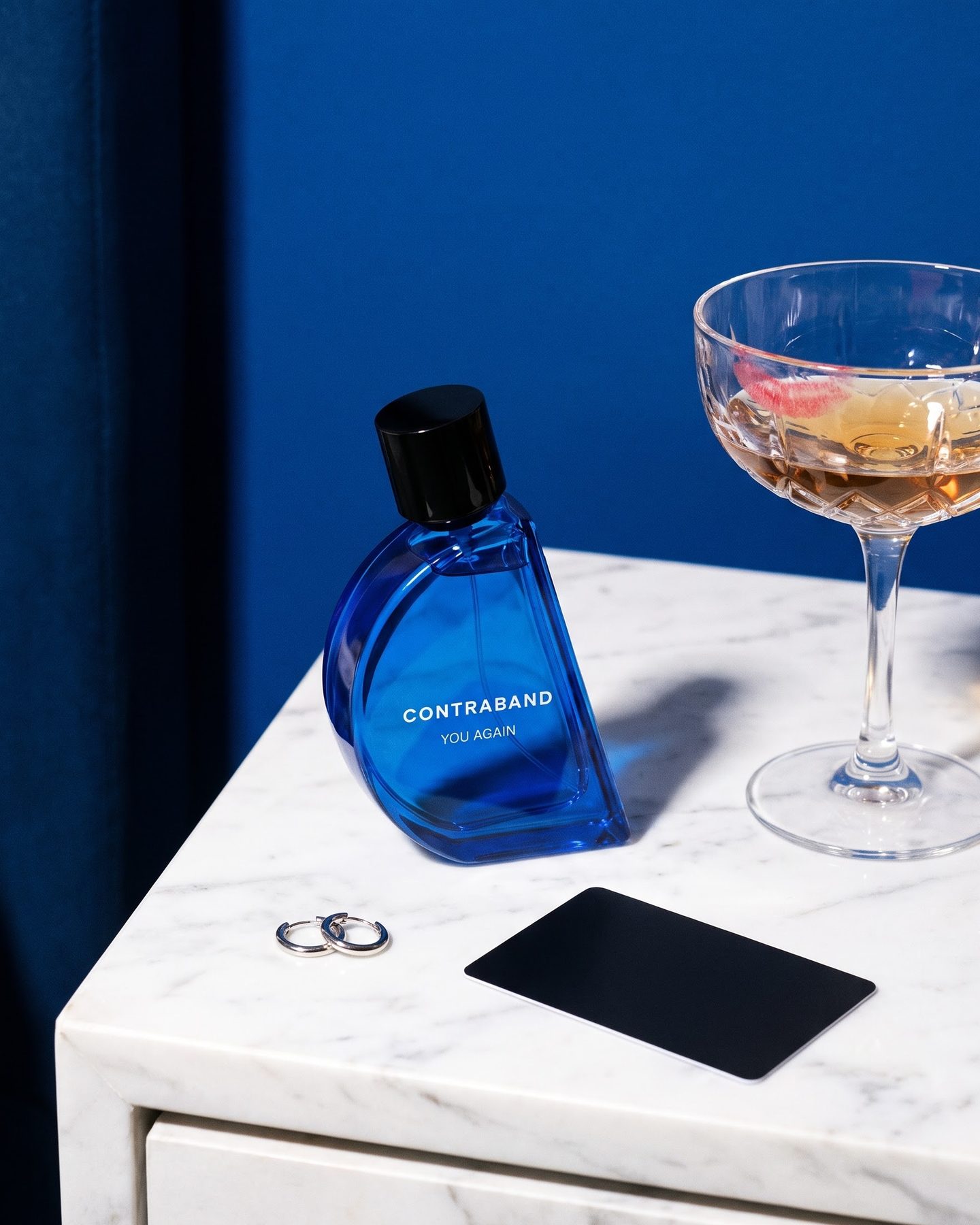 Still life: the Contraband You Again bottle on a white marble nightstand against a cobalt wall, beside a silver hoop earring, a black key card and a lipstick-marked coupe.