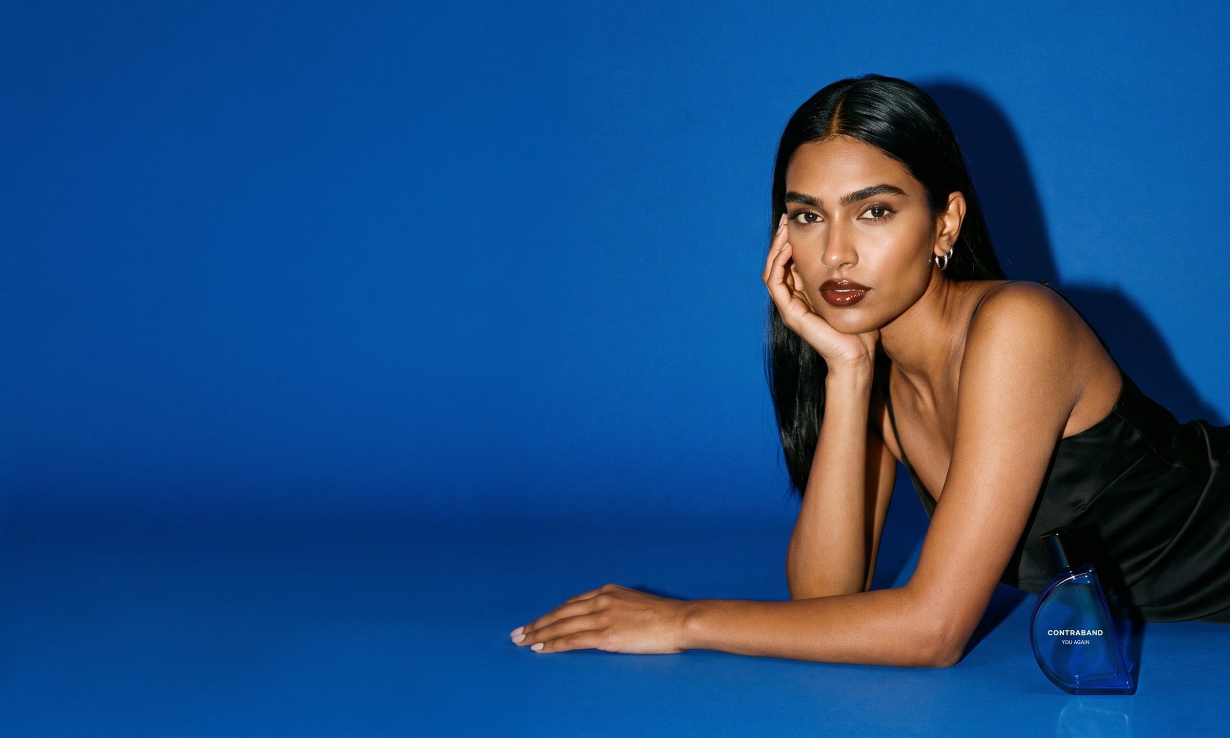 A woman in a black satin slip lies on a seamless cobalt-blue set, chin in hand, looking into the lens, with a cobalt Contraband You Again bottle beside her elbow.