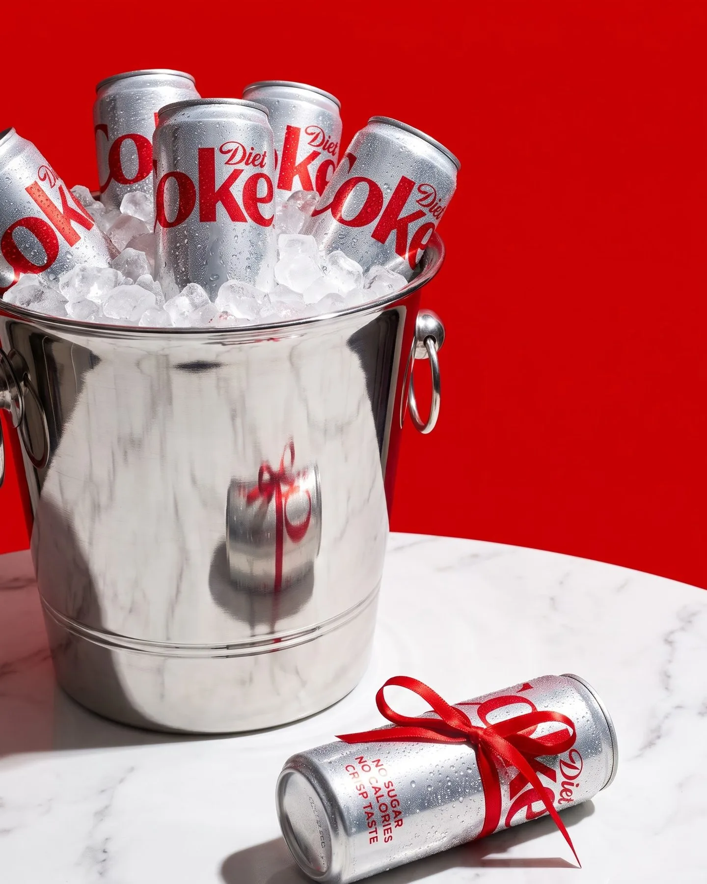 Cans in a silver ice bucket on white marble against a red wall, one can tied with a red ribbon.
