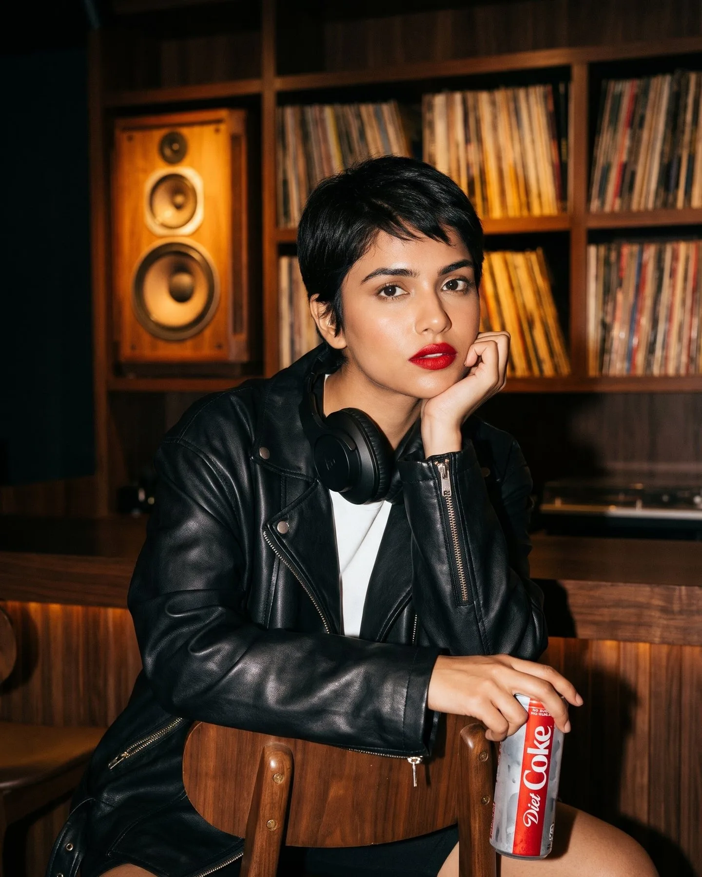 A woman with a pixie cut and leather jacket leans on a walnut bar in a vinyl listening lounge, can in hand.