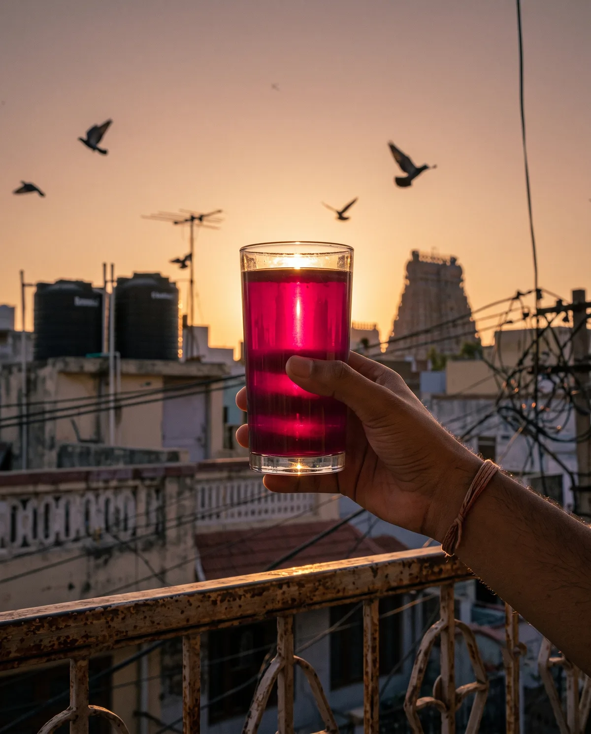 A glass of drink held against a balcony railing at dusk, city rooftops behind